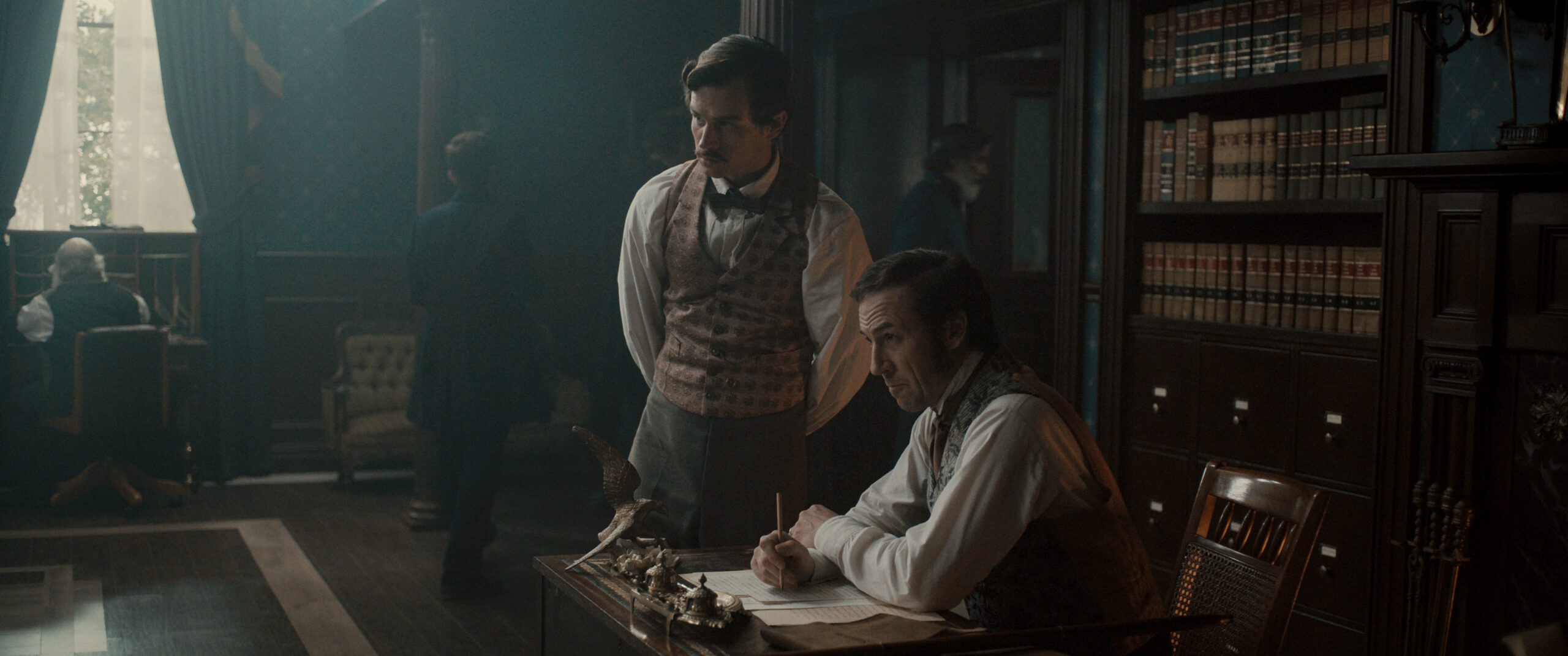 Still from Manhunt Season 1 Episode 6 of Brandon Flynn and Tobias Menzies pictured from left to right. Menzies is seated at a desk and Flynn is standing next to him.