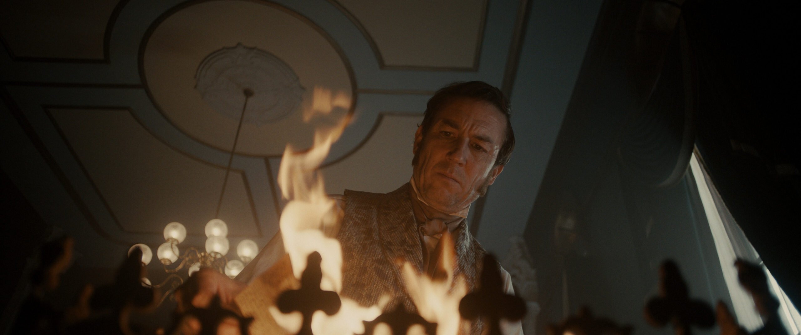 Still from Manhunt Season 1 Episode 6 of Tobias Menzies. Photo is angled from below Menzies as he looks at a flame below him.