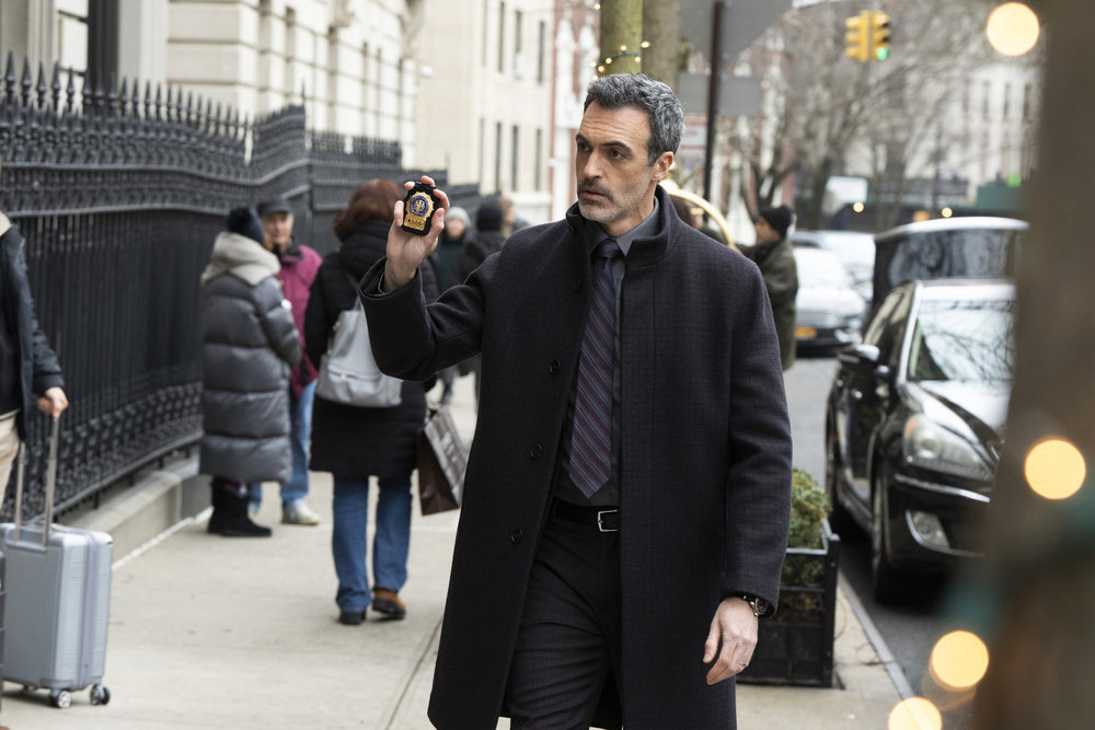 Law & Order Season 23 Episode 9 - Reid Scott as Det. Vincent Riley stands on a Manhattan sidewalk and holds up his police badge.