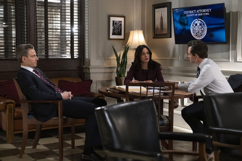 Law & Order Season 23 Episode 9 - Tony Goldwyn as DA Nicholas Baxter sits with Odelya Halevi as ADA Samantha Maroun and Hugh Dancy as ADA Nolan Price discussing the case.