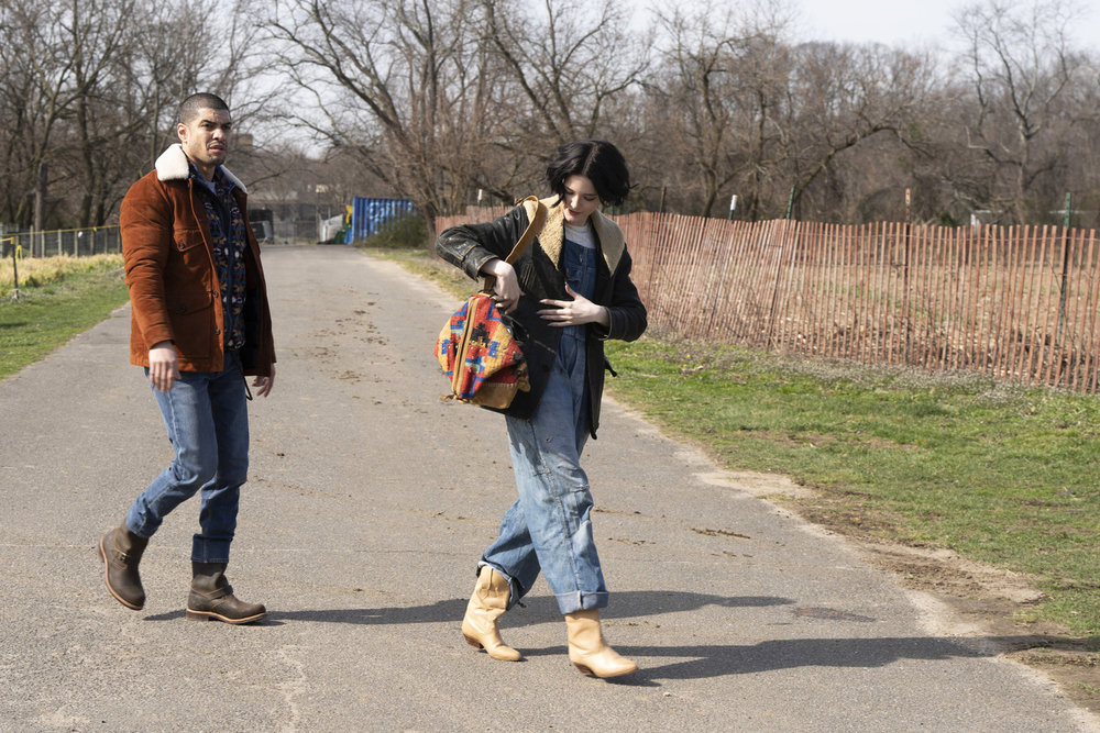 Law & Order: Organized Crime Season 4 Episode 9 - Rick Gonzalez as Det. Bobby Reyes walks across a country road with Ainsley Seiger as Det. Jet Slootmaekers, who is digging into a bag over her shoulder.