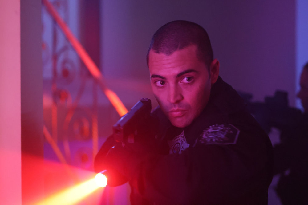 Chicago P.D. Season 11 Episode 10 - Benjamin Levy Aguilar as Dante Torres stands at attention with his flashlight in one hand crossed over to be positioned just below his other hand which is yielding his gun.