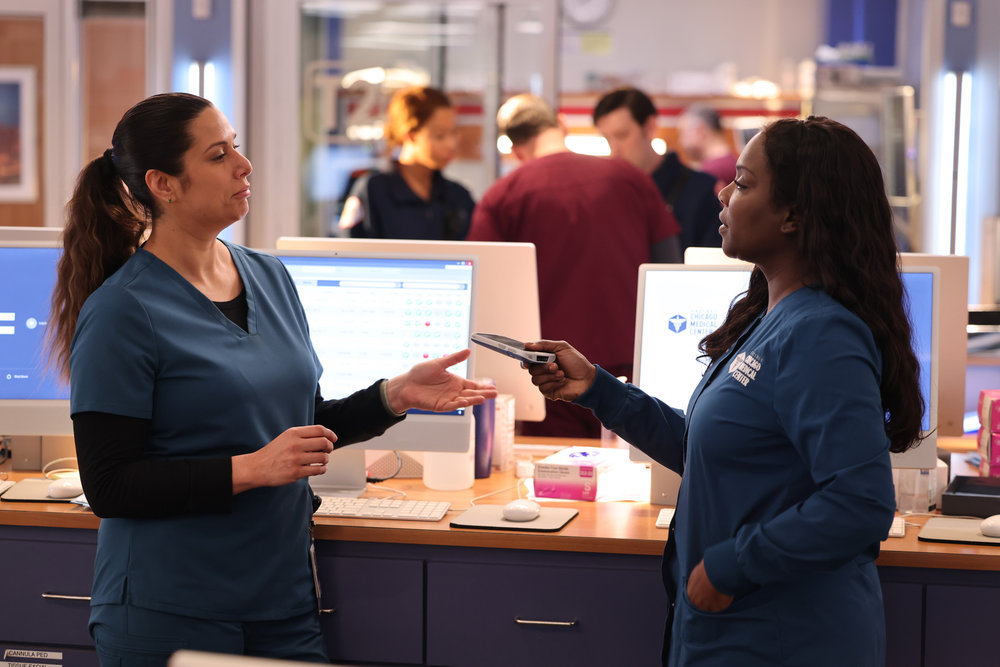 Chicago Med Season 9 Episode 10 - Lorena Diaz as Nurse Doris stands with Marlyne Barrett as Maggie Lockwood as she hands her a work phone.