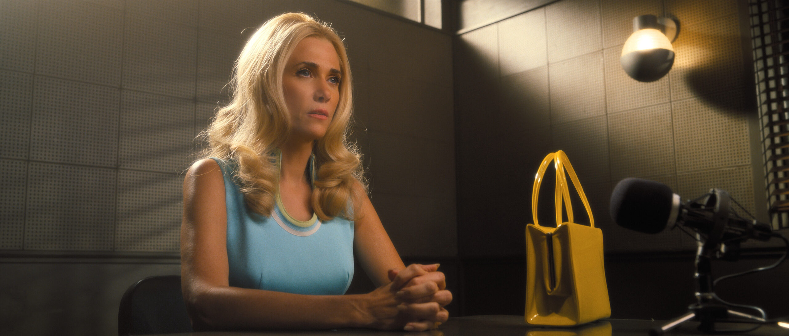 Maxine wears a powder blue sleeveless dress with her blonde hair cascading down her shoulders. She sits in a police station interrogation room on Palm Royale Season 1 Episode 7, "Maxine Bags a Prince."