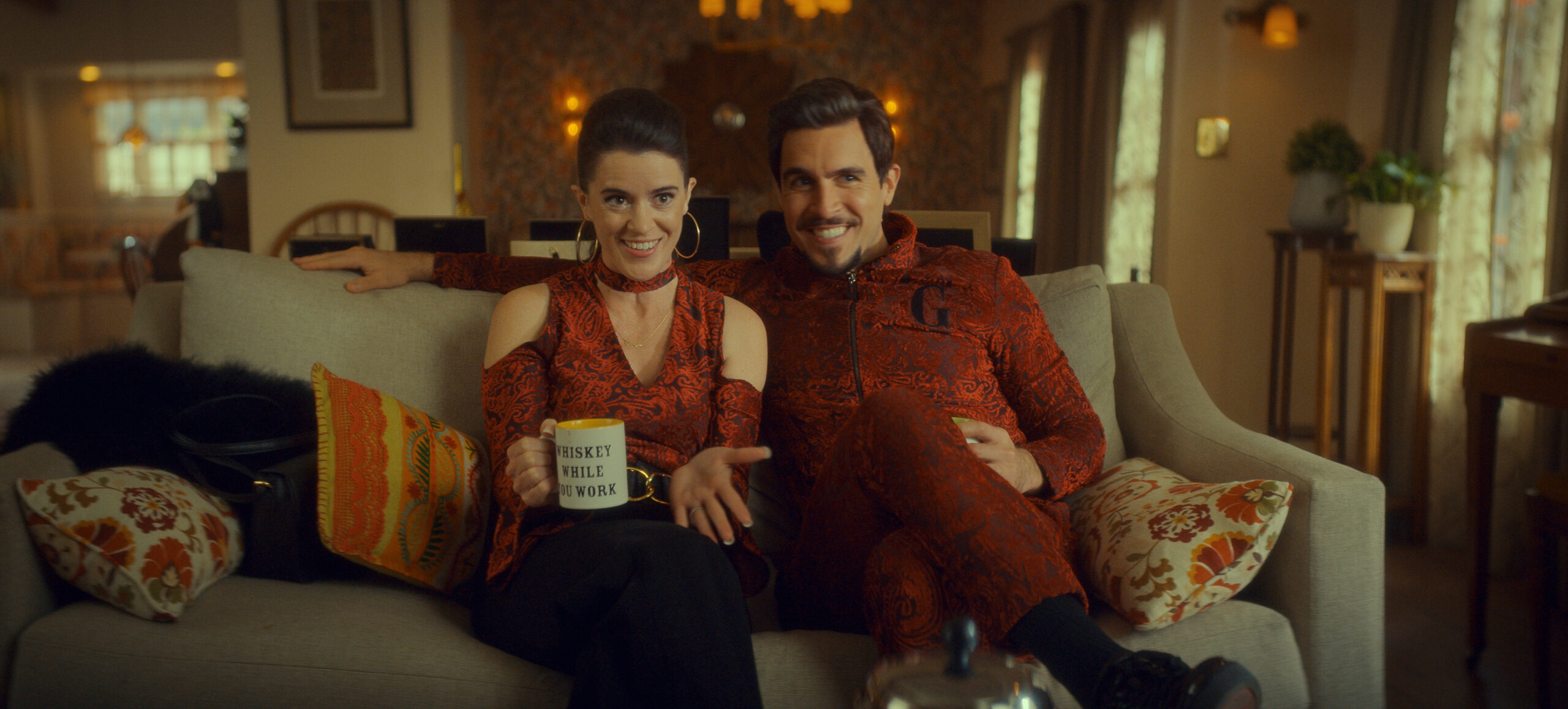 Still. from The Big Door Prize Season 2 Episode 9 of Mary Holland and Josh Segarra sitting on a couch from left to right wearing clothes with a matching red pattern.
