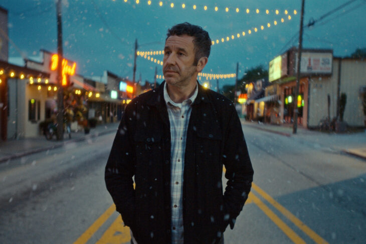 Still from The Big Door Prize Season 2 Episode 10 of Chris O’Dowd walking down an street in snowy weather wearing a black jacket and a blue and white plaid shirt.