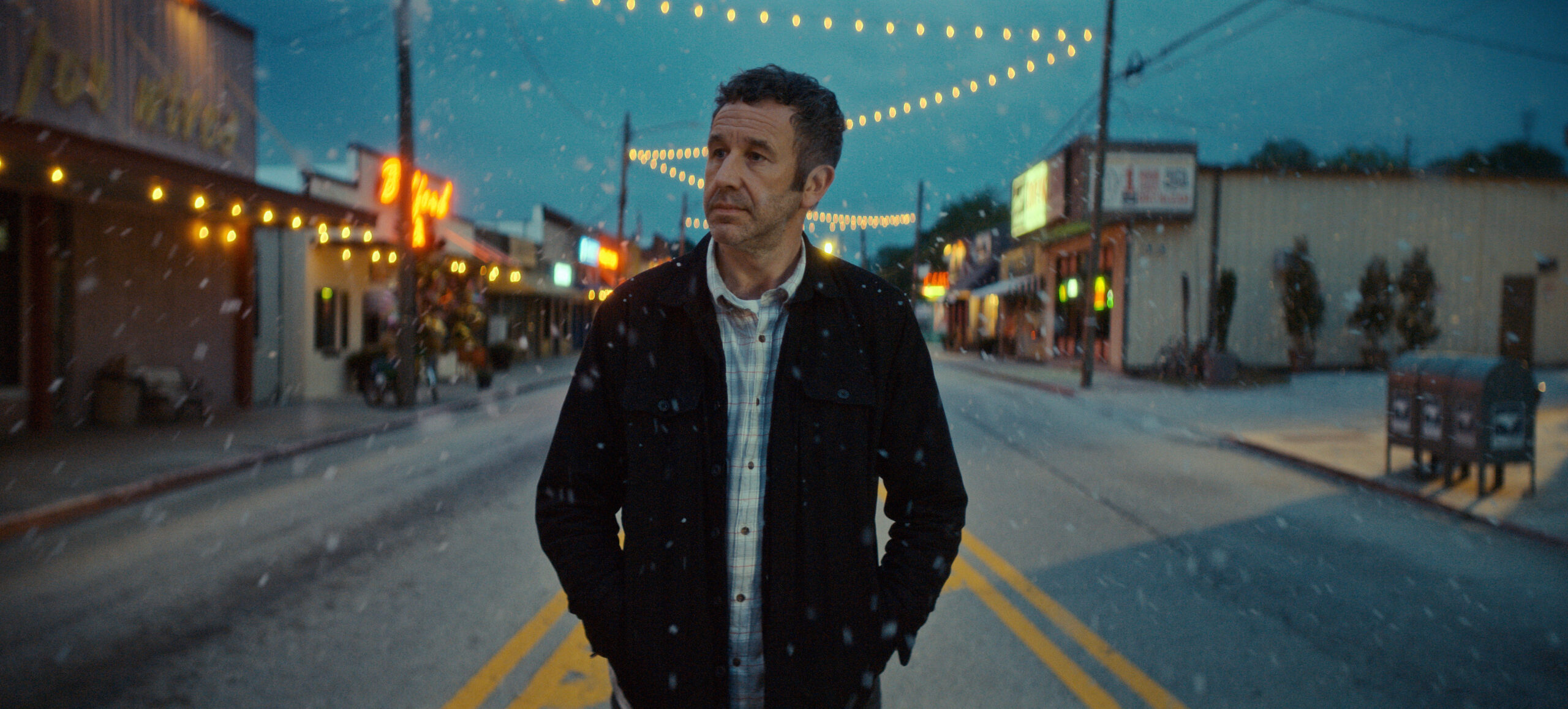 Still from The Big Door Prize Season 2 Episode 10 of Chris O’Dowd walking down an street in snowy weather wearing a black jacket and a blue and white plaid shirt.