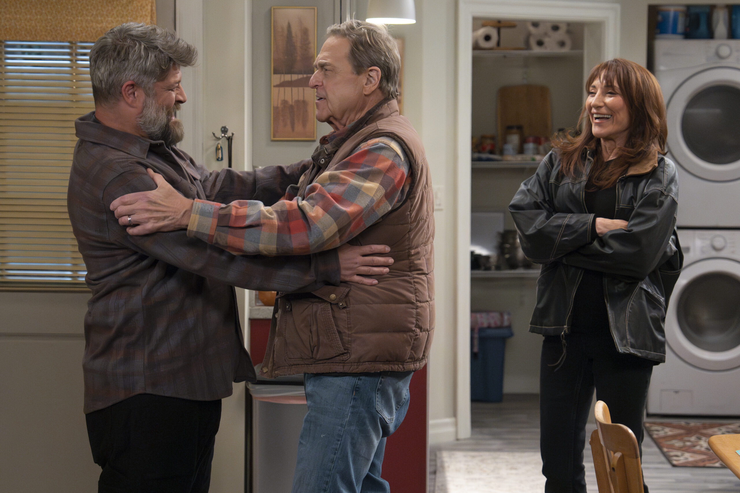 JAY R. FERGUSON, JOHN GOODMAN, KATEY SAGAL | Tell-Tale TV The Conners Season 6 Episode 11 JAY R. FERGUSON smiles with JOHN GOODMAN, as their arms are around each other while KATEY SAGAL stands nearby laughing.