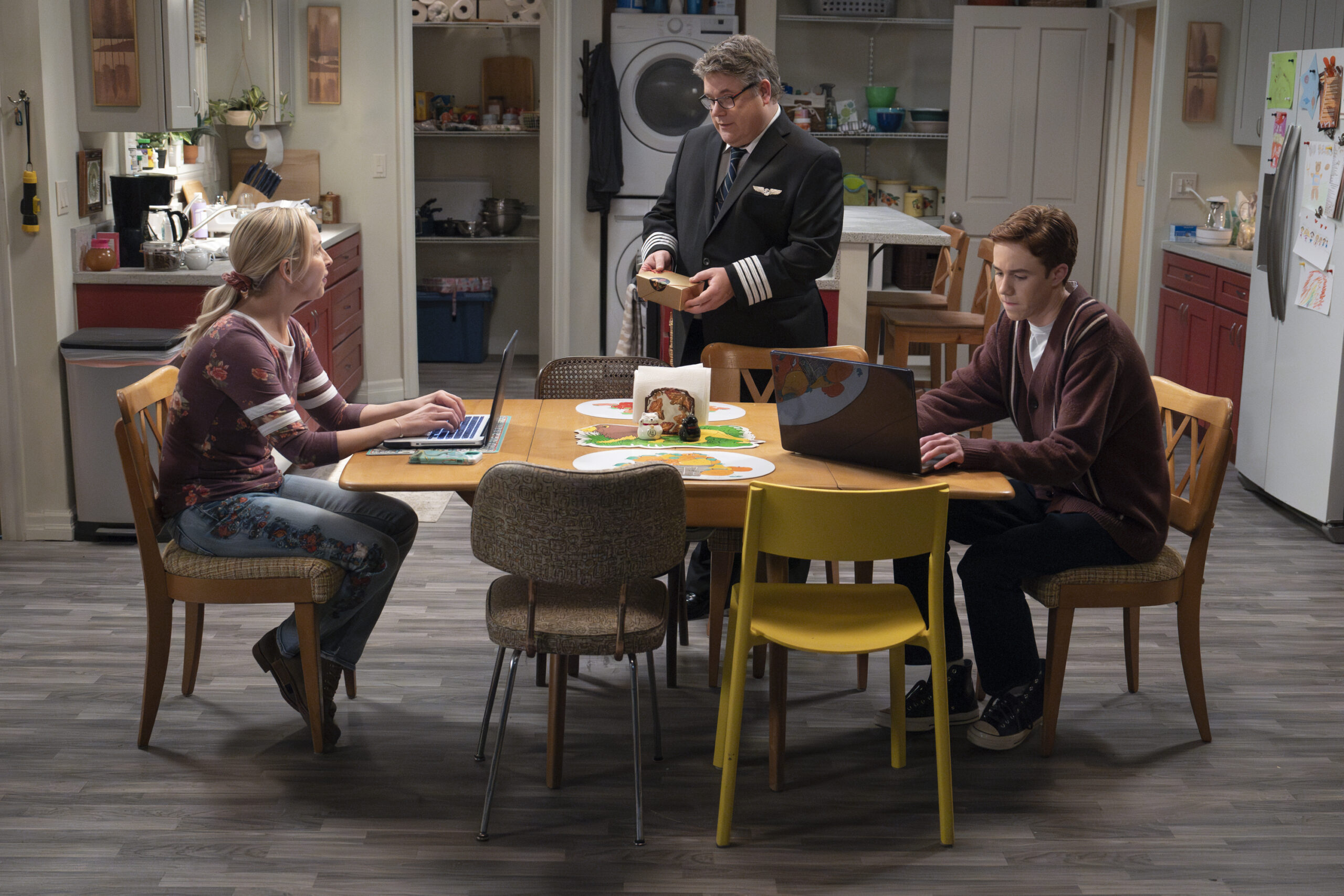 LECY GORANSON, SEAN ASTIN, AMES MCNAMARA | Tell-Tale TV The Conners Season 6 Episode 11 LECY GORANSON sits at her kitchen table typing while SEAN ASTIN stands nearby with a treat and AMES MCNAMARA sits across from her also typing.