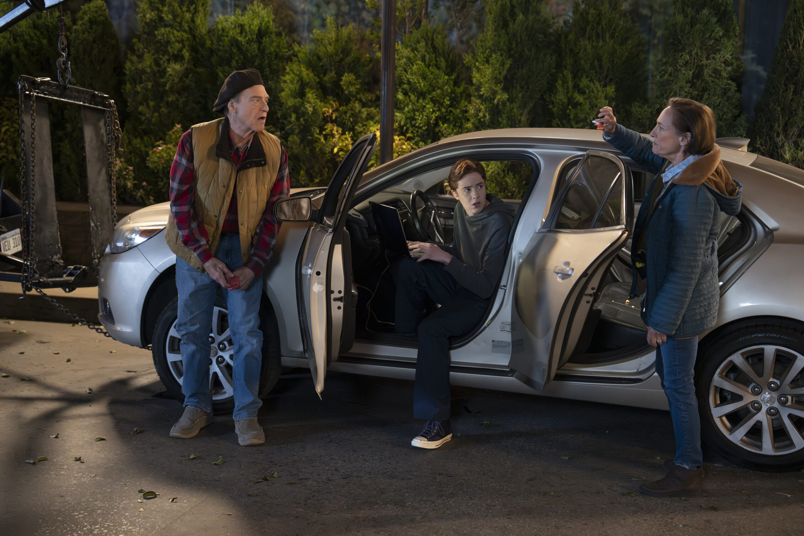 JOHN GOODMAN, AMES MCNAMARA, LAURIE METCALF | Tell-Tale TV The Conners Season 6 Episode 13 - JOHN GOODMAN leans on the front of AMES MCNAMARA car, while he sits half in, half out of the drivers seat and LAURIE METCALF stands at the back of the car.