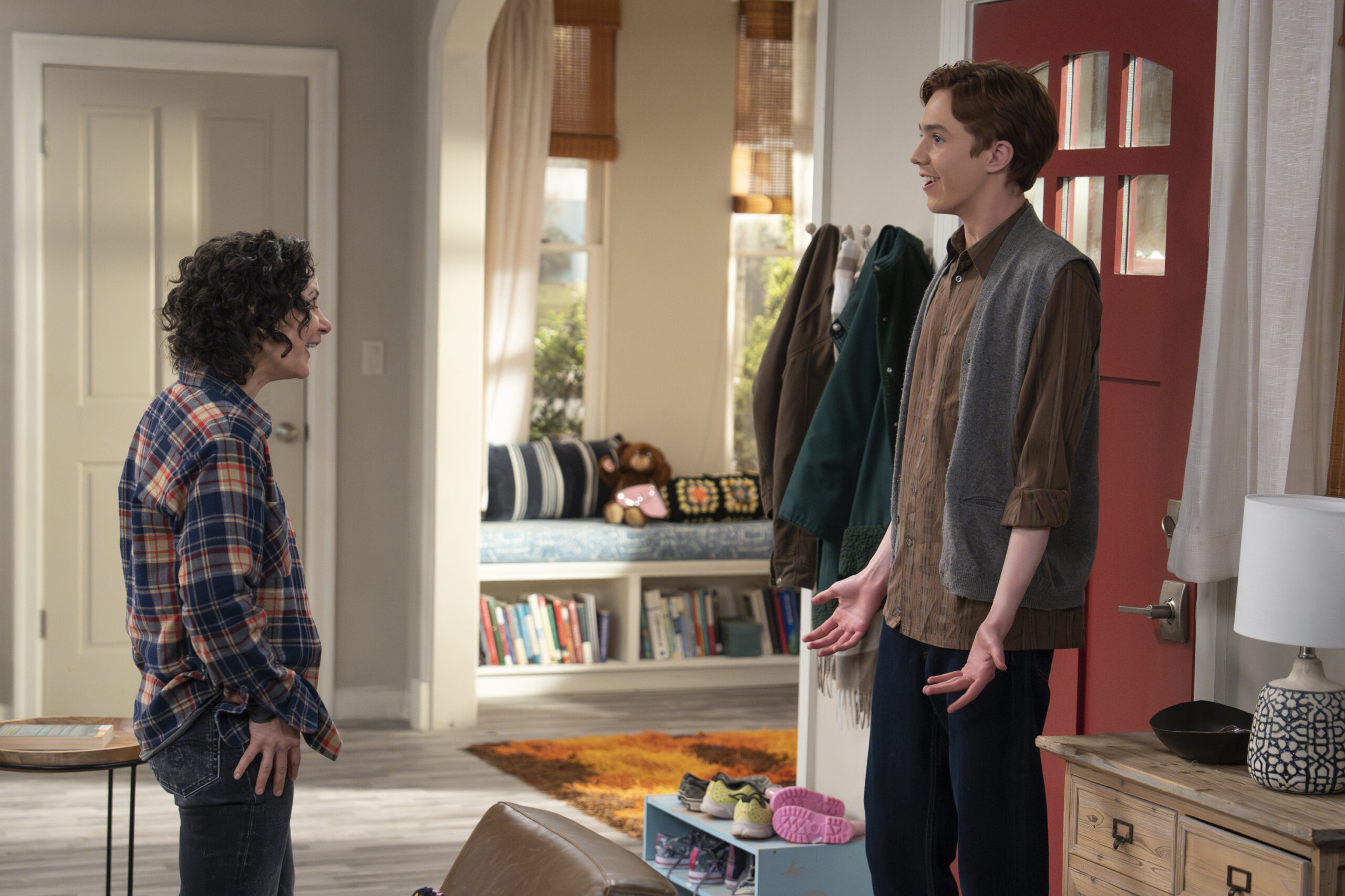 SARA GILBERT, AMES MCNAMARA | Tell-Tale TV The Conners Season 6 Episode 13 - SARA GILBERT and AMES MCNAMARA stand in their living room with smiles on their faces, talking.