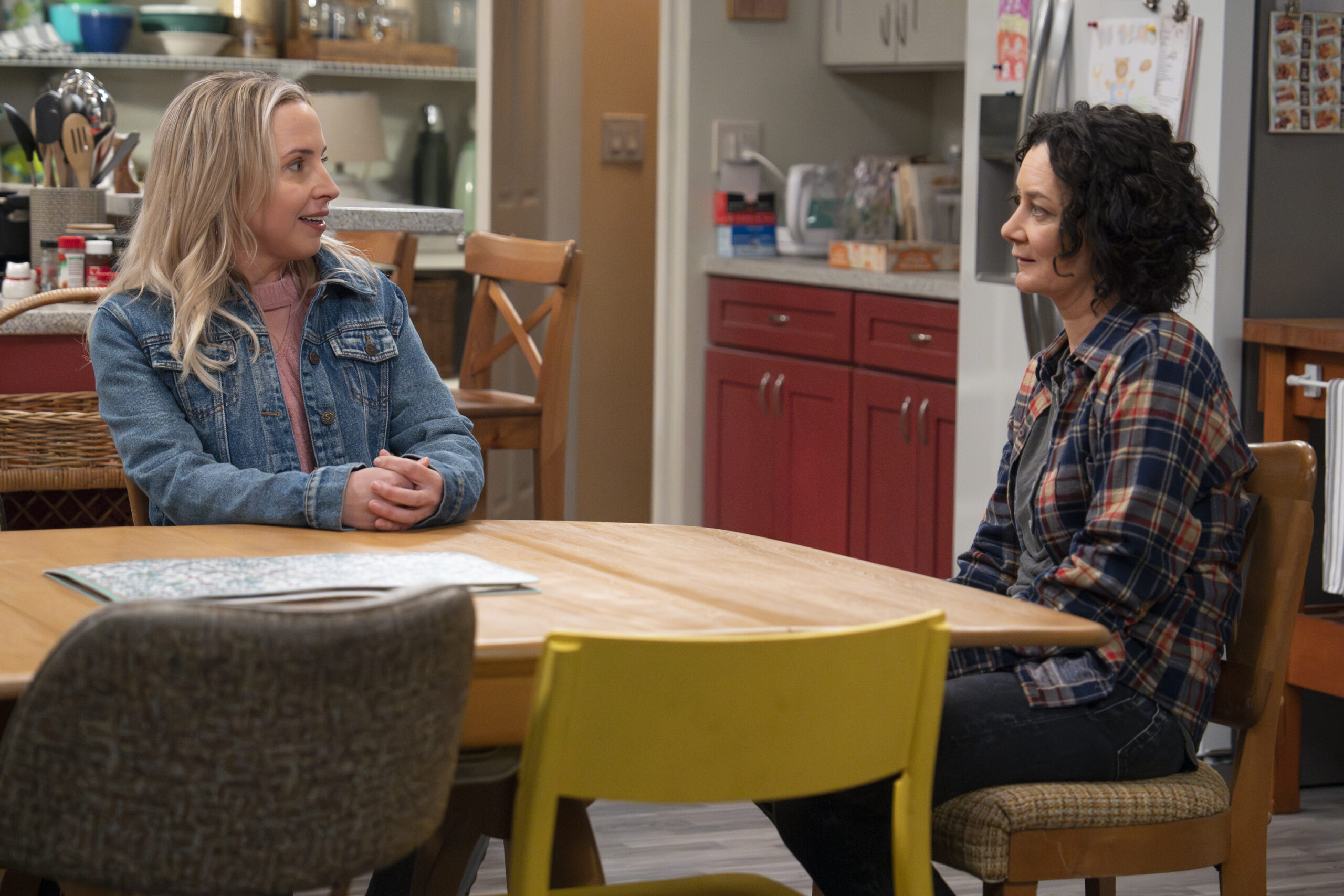 LECY GORANSON, SARA GILBERT | Tell-Tale TV The Conners Season 6 Episode 13 - LECY GORANSON and SARA GILBERT sit at their kitchen table, talking.