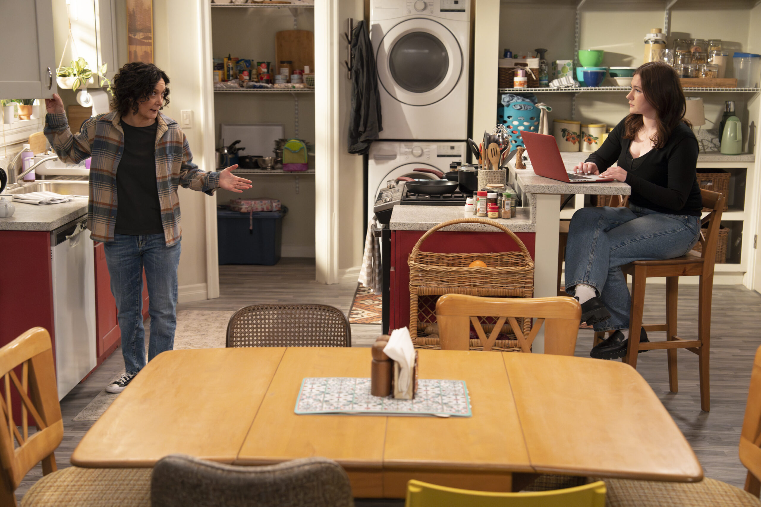 SARA GILBERT, EMMA KENNEY | Tell-Tale TV The Conners Season 6 Episode 13 - SARA GILBERT stands near EMMA KENNEY who is working on her laptop at the kitchen counter.