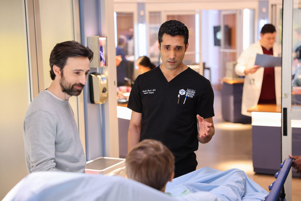 Chicago Med Season 9 Episode 12 - Josh Zuckerman as Bill Storch stands smiling at a child in the bed while Dominic Rains as Dr. Crockett Marcel explains something.