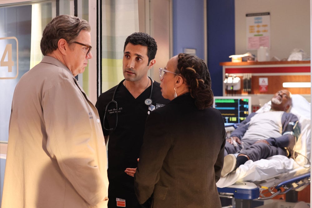 Chicago Med Season 9 Episode 12 - Oliver Platt as Dr. Daniel Charles looks at S. Epatha Merkerson as Sharon Goodwin with concern as Dominic Rains as Dr. Crockett Marcel explains what is wrong with the patient.