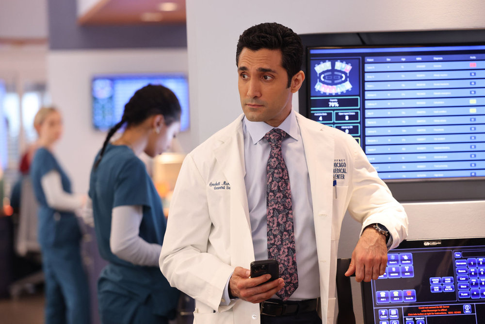 Chicago Med Season 9 Episode 12 - Dominic Rains as Dr. Crockett Marcel stands looking off to the side, leaning on a computer moniter with his phone out in one hand.
