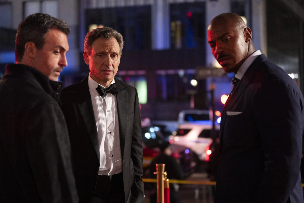 Law & Order Season 23 Episode 13 - Reid Scott as Det. Vincent Riley and Mehcad Brooks as Det. Jalen Shaw look over their shoulders at the murder victim while Tony Goldwyn as DA Nicholas Baxter stands nearby in shock.