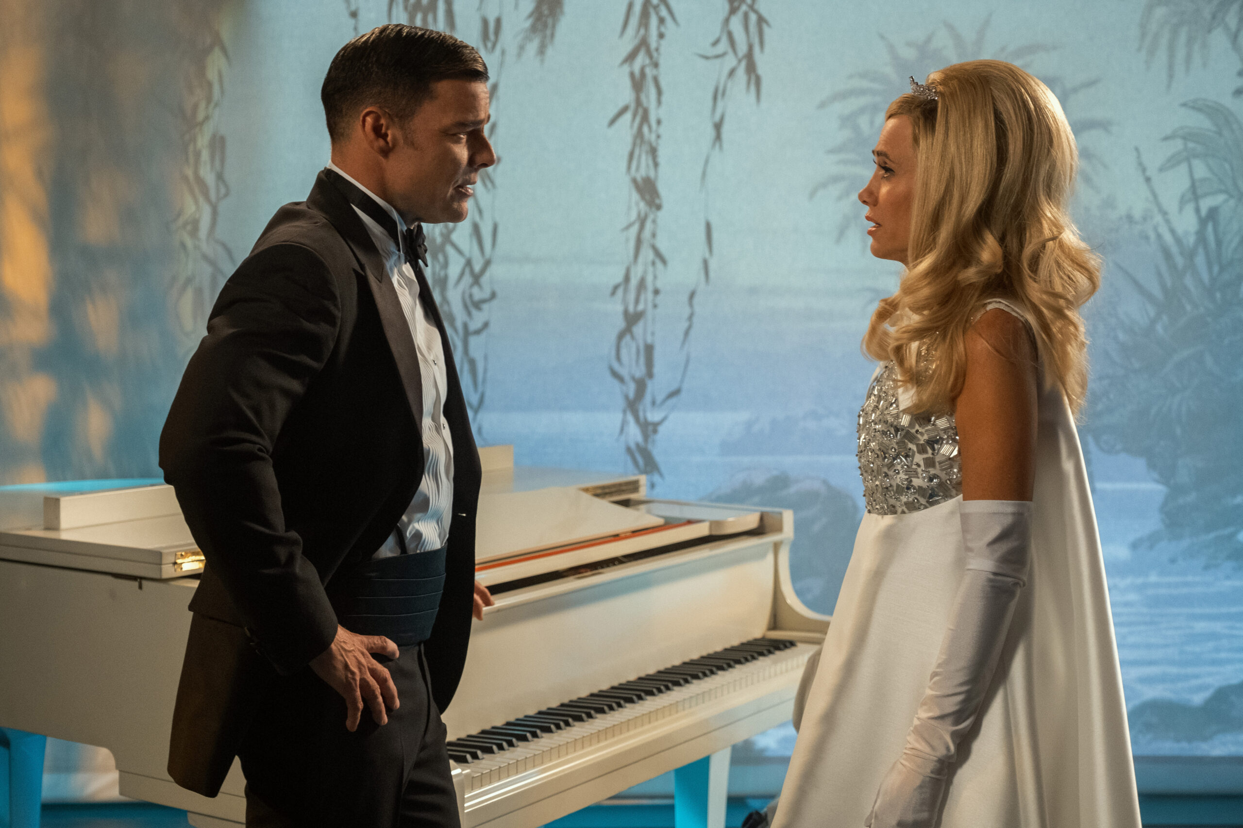 Robert wears a black suit jacket with matching dress pants and a white dress shirt with a black bow tie. He stands across from Maxine, who wears a short-sleeved floor-length white dress with matching white gloves and a glittering tiara. They stand in front of a white piano on a stage on Palm Royale Season 1 Episode 10, "Maxine Throws a Party."