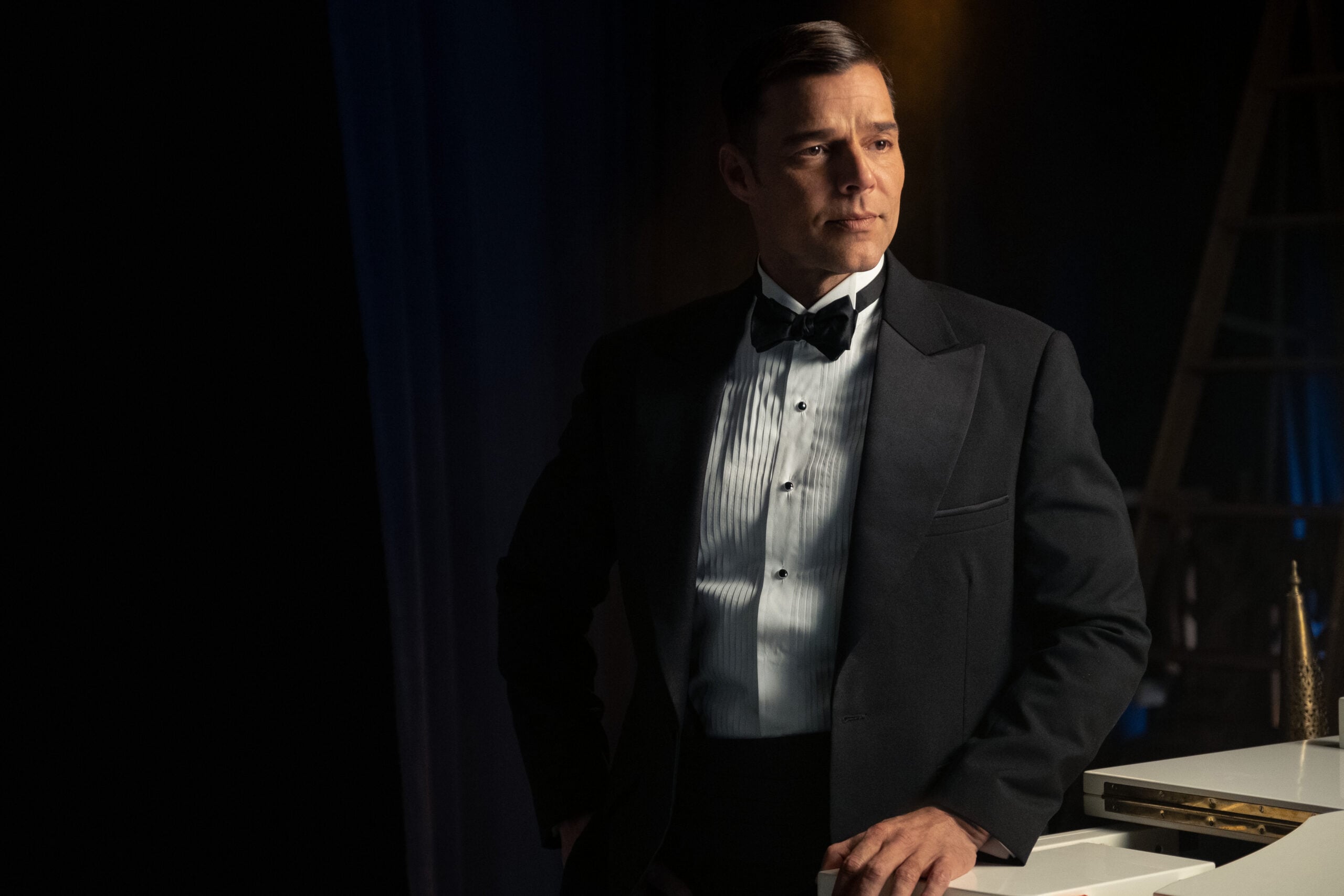 Robert wears a white dress shirt with a black bow tie, a black suit jacket, and black dress pants. He stands in a dimly lit room while looking off to the side in a pensive manner on Palm Royale Season 1 Episode 10, "Maxine Throws a Party."