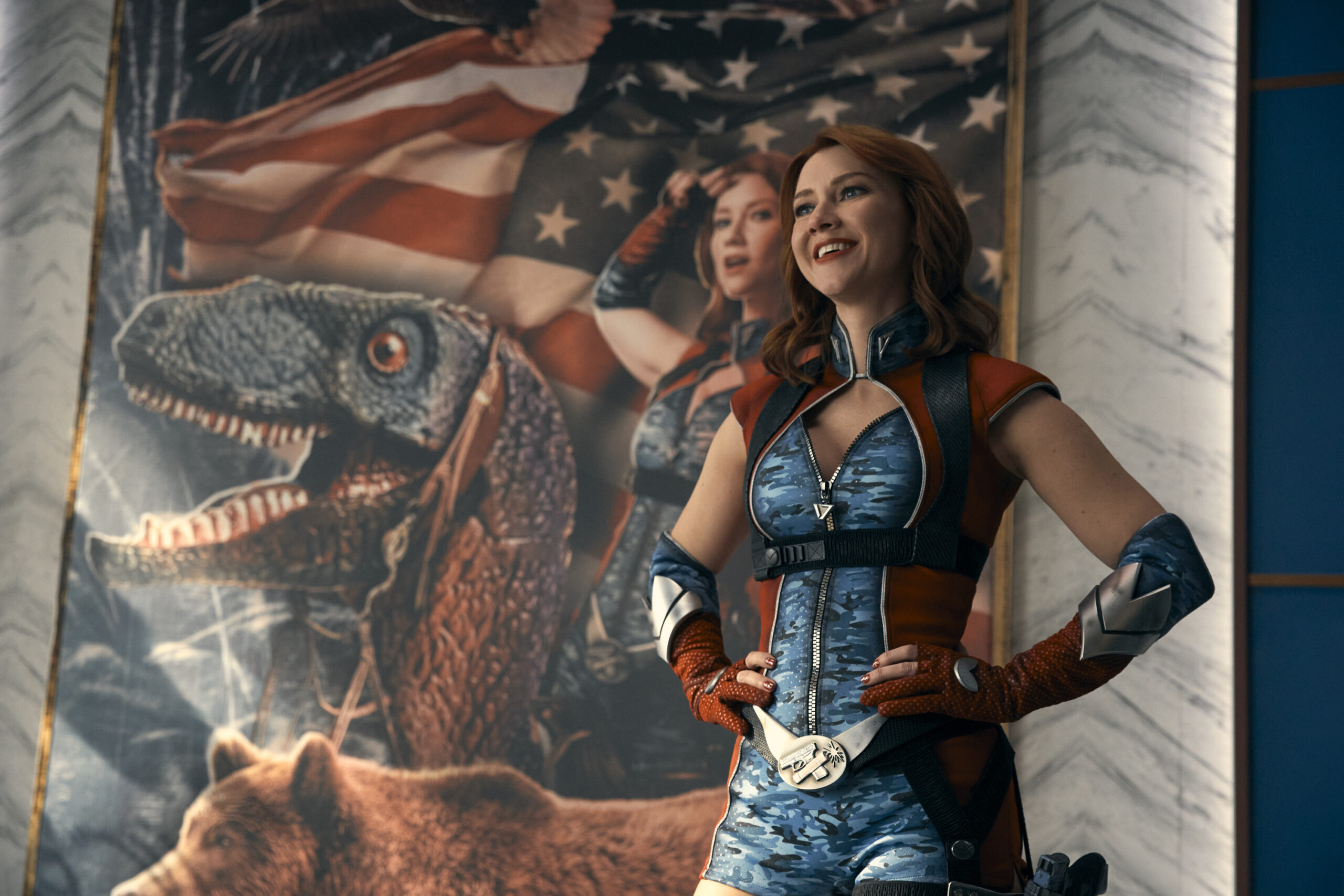Still from The Boys Season 4 Episode 3 of a Supe in full costume standing in front of a portrait of herself with a dinosaur, bear and American flag on it.