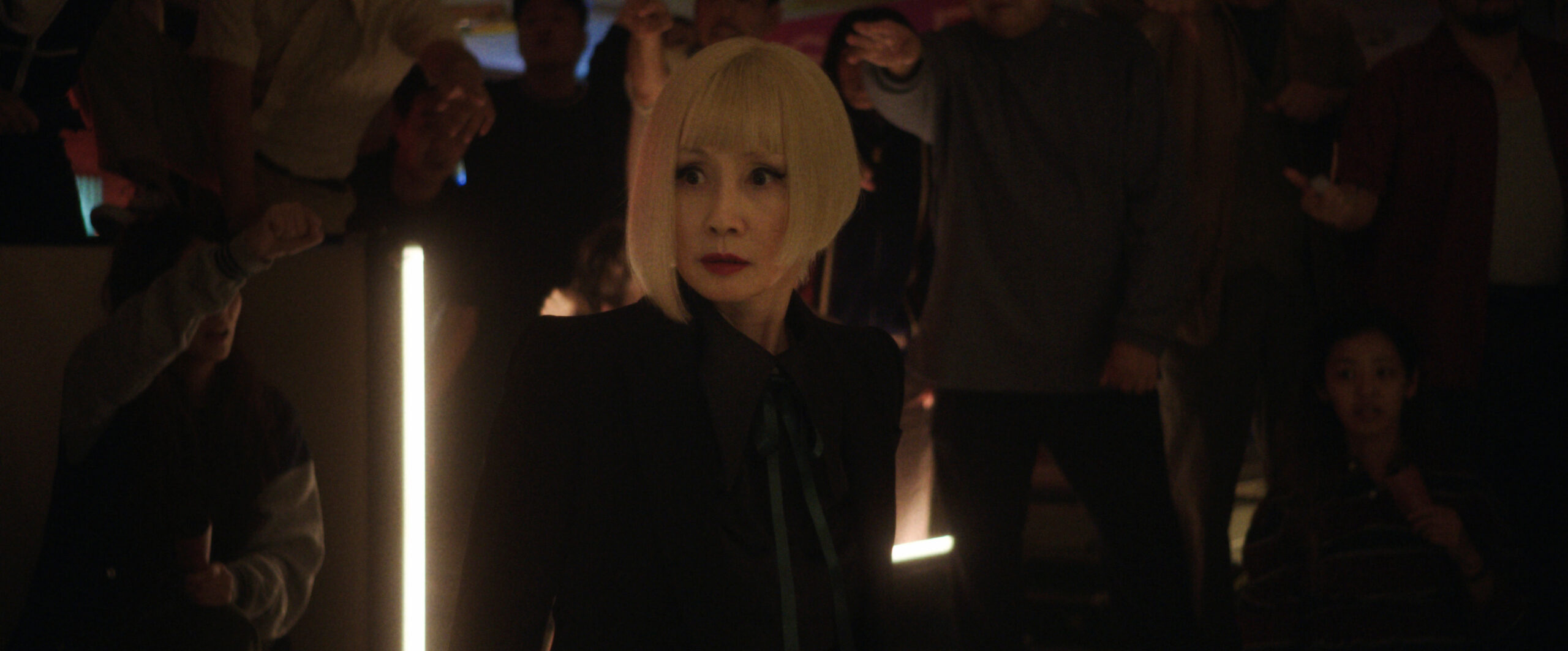 Sunny Season 1 Episode 3 Mmmm Hinoki | Tell-Tale TV Hime looks at someone off-screen in shock while standing in a dimly lit room that hosts robot fights on Sunny Season 1 Episode 3, "Mmmm Hinoki."