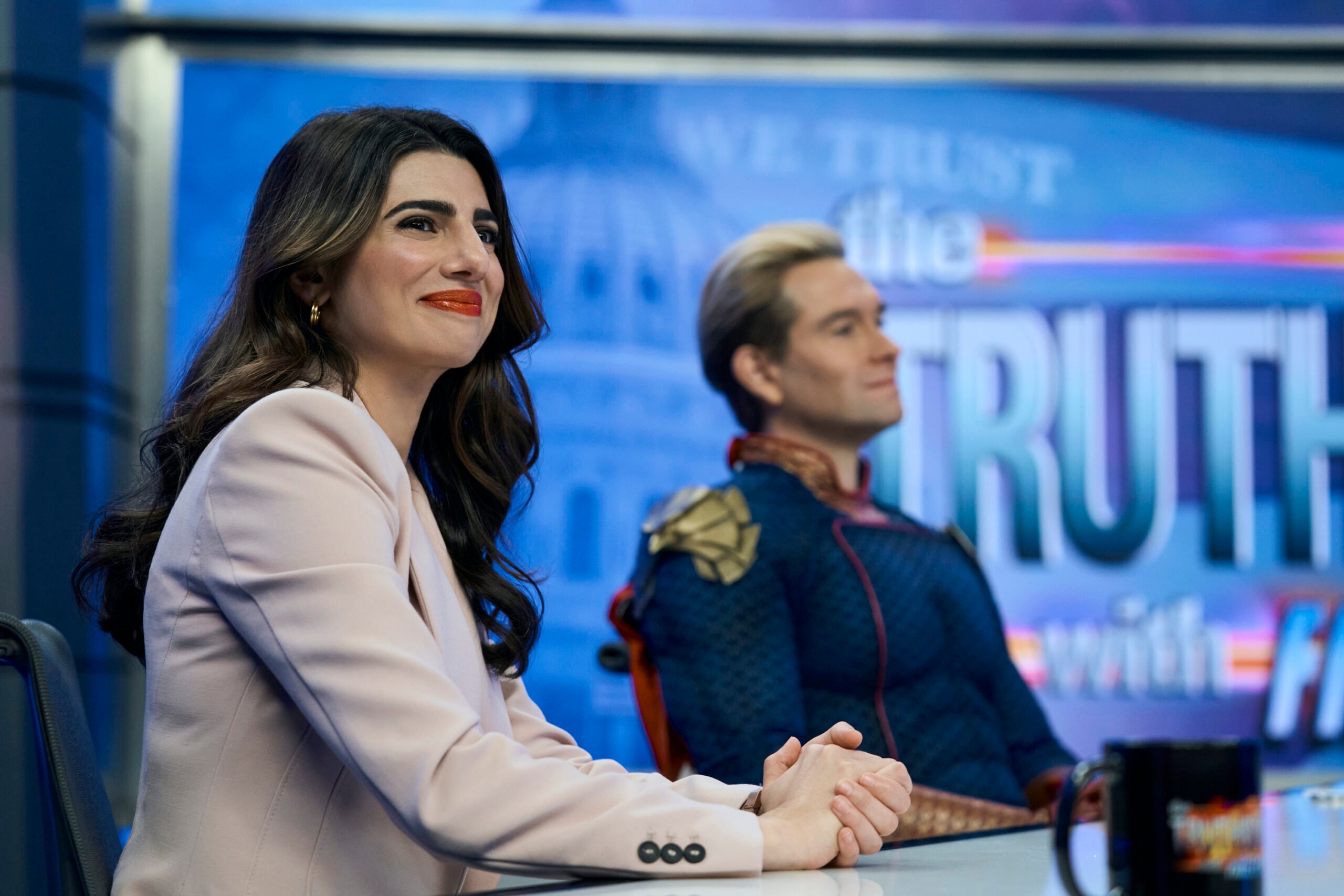 The Boys Season 4 Episode 8 - Claudia Doumit and Antony Starr | Tell-Tale TV Still from The Boys Season 4 Episode 8 of Claudia Doumit as Victoria Neuman and Antony STarr as Homelander pictured from left to right sitting at table in a TV studio.