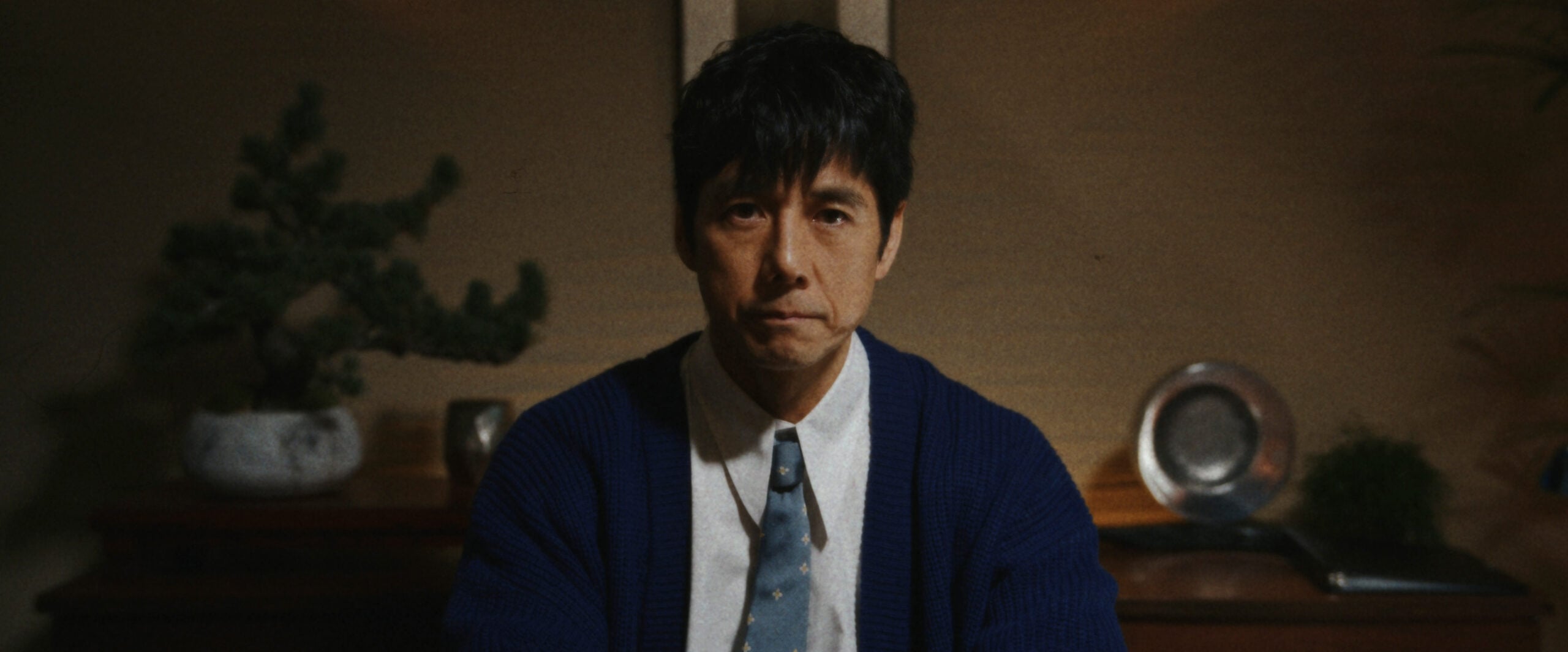 Masa wears a button-down shirt with a tie and a navy blue cardigan while sitting in a dimly lit office on Sunny Season 1 Episode 9, "Who's in the Box?"