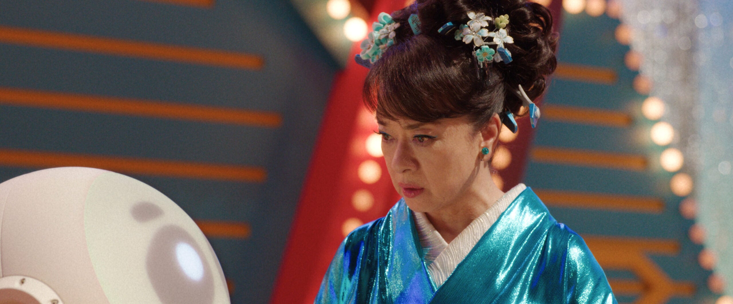 Noriko wears a teal satin kimono with teal and white accessories in her hair. She stands on a Japanese gameshow stage on Sunny Season 1 Episode 9, "Who's in the Box?"