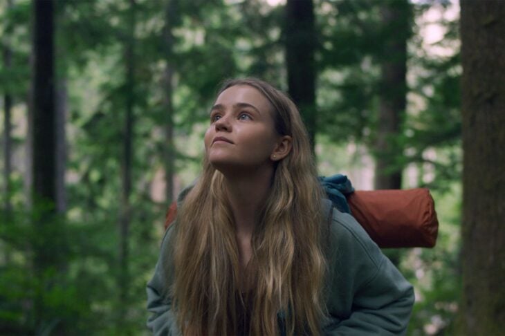 A still from the Netflix series Penelope, featuring a teenage blonde white girl who walks through the woods with a backpack and sleeping bag on her back.