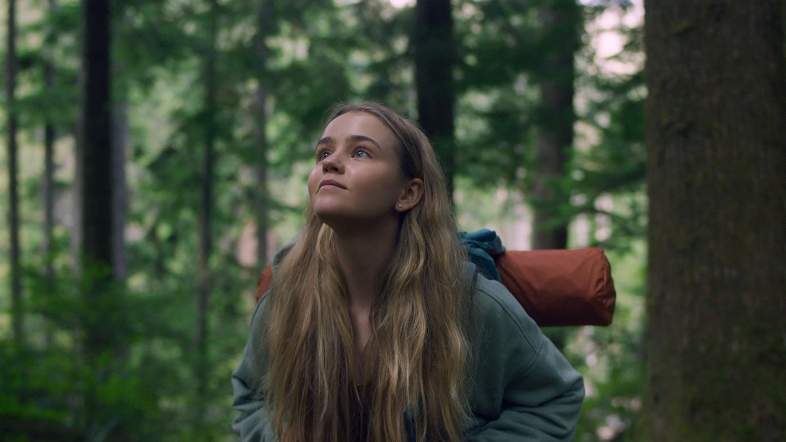 A still from the Netflix series Penelope, featuring a teenage blonde white girl who walks through the woods with a backpack and sleeping bag on her back.