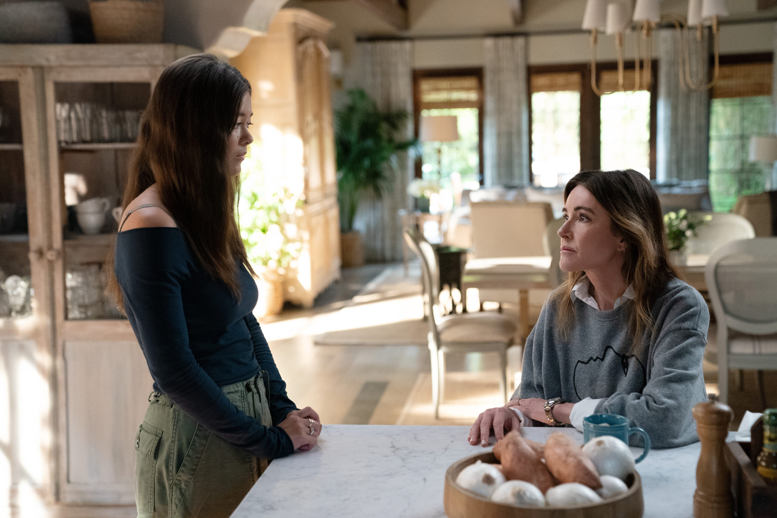 Alice stands in a brightly lit kitchen while Liz looks up at her, sitting at the kitchen island, on Shrinking Season 2 Episode 5, "Honesty Era."