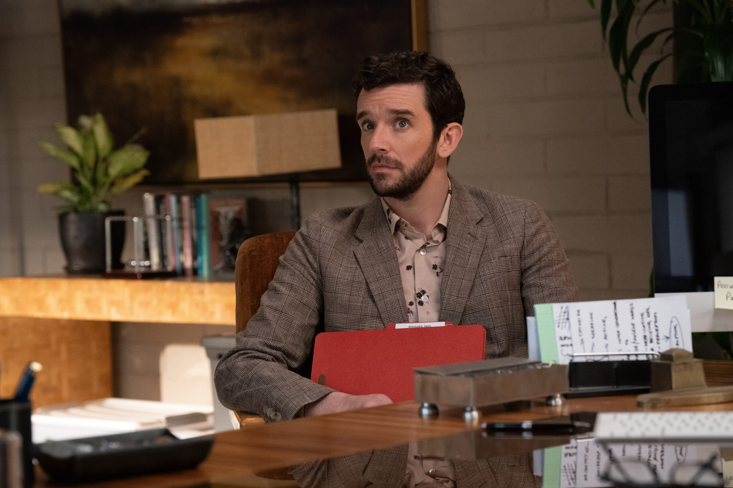 Brian sits at a desk in an office while holding a folder and listening to someone intently on Shrinking Season 2 Episode 5, "Honesty Era."