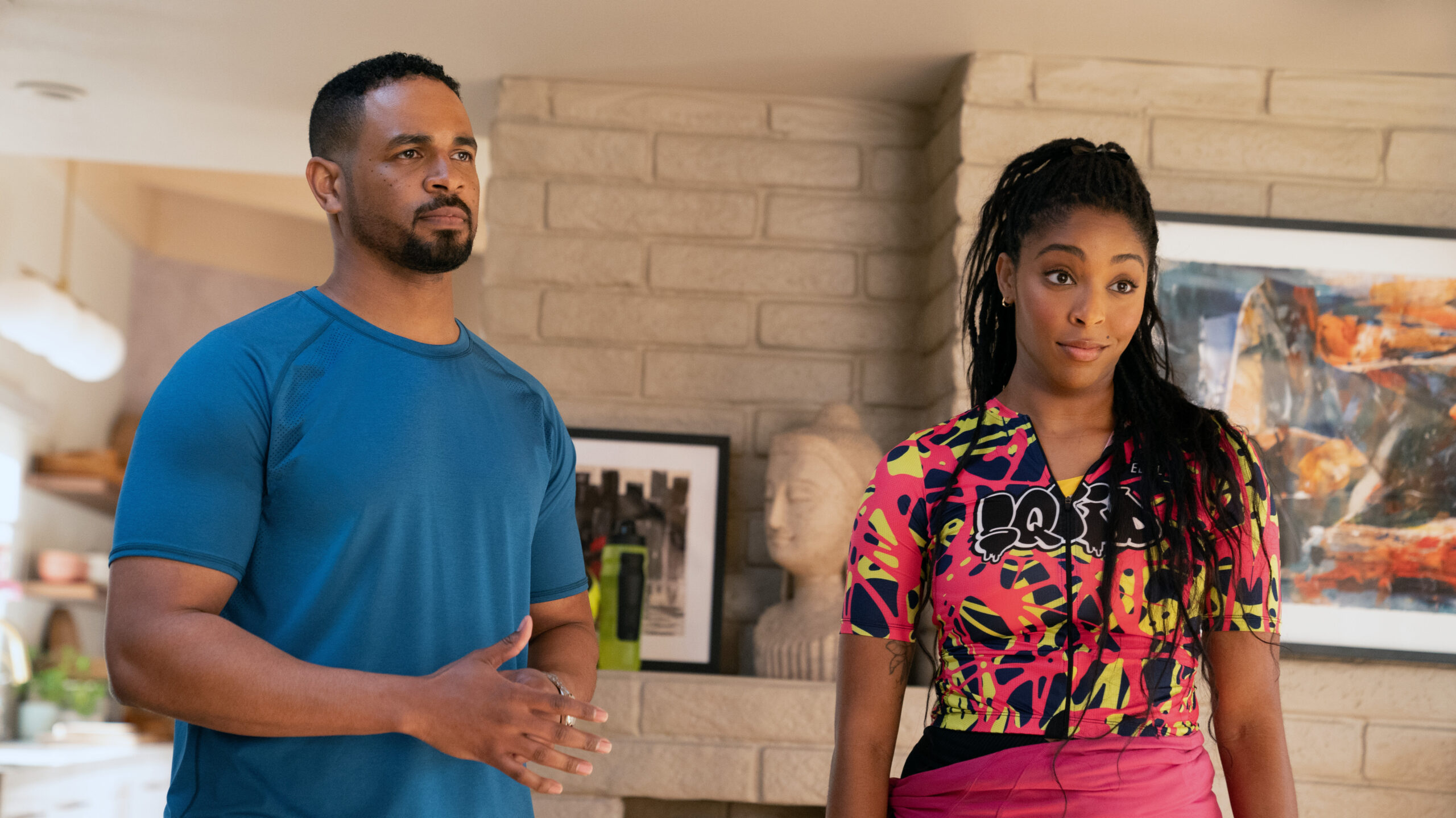 Shrinking Season 2 Episode 7 Get in the Sea | Tell-Tale TV Derrick and Gaby stand in the latter's brightly lit living room while wearing biking gear on Shrinking Season 2 Episode 7, "Get in the Sea."